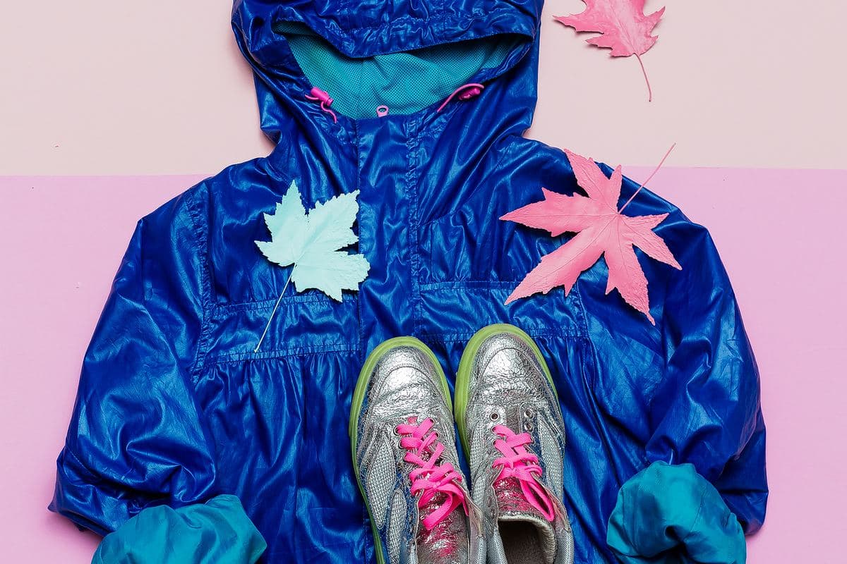 Un impermeable blau brillant estès en zenital sobre fons rosa, amb unes sabatilles platejades i fulles pintades de rosa